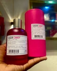 Two perfume bottles labeled 'Sugar Candy' (one dark red glass bottle, one bright pink spray can), held in a hand in front of a blurred background with colorful lights, including a barcode on the labels.