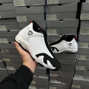 Person holding a white and black athletic sneaker with a Jumpman logo, in front of stacked shoeboxes in a store.